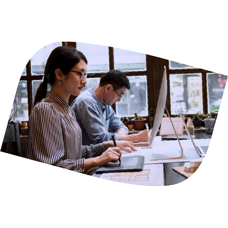 Woman in foreground sit and desk and draws on a table while man works beside her